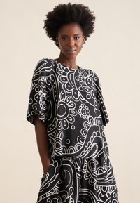 Top a motivi bianchi e neri con maniche corte, caratterizzato da disegni floreali e paisley. La texture del tessuto appare liscia e leggera.
