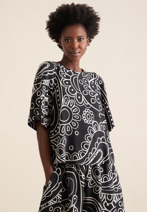 Top a motivi bianchi e neri con maniche corte, caratterizzato da disegni floreali e paisley. La texture del tessuto appare liscia e leggera.