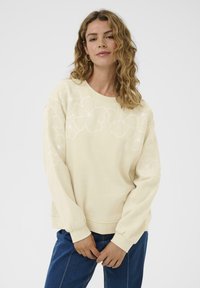 Femme avec des cheveux ondulés portant un sweat-shirt crème avec des broderies florales sur la poitrine et les manches, associé à un jean bleu, devant un fond blanc.