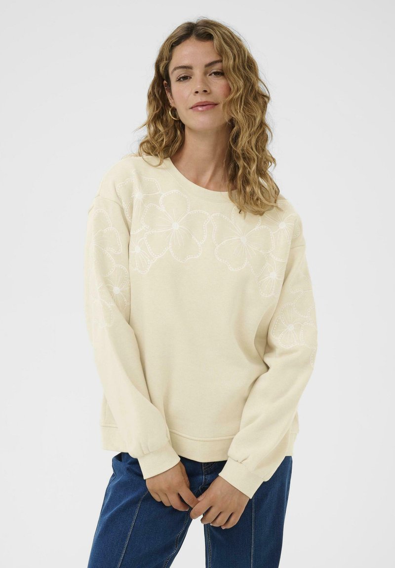 Femme avec des cheveux ondulés portant un sweat-shirt crème avec des broderies florales sur la poitrine et les manches, associé à un jean bleu, devant un fond blanc.
