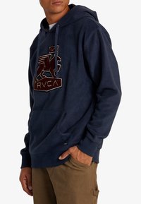 Sudadera con capucha de forro polar en color marino, con un bolsillo tipo canguro, que presenta un logotipo RVCA en relieve en marrón y blanco y una capucha con cordón. Textura suave, corte relajado.