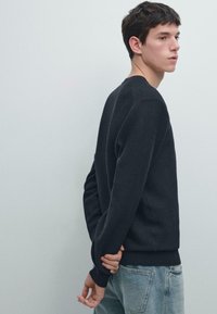 Maglione nero con trama, scollo rotondo e maniche lunghe, abbinato a jeans azzurri. Il maglione presenta orlo e polsini a costine.