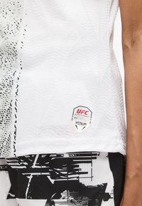 Λευκό αθλητικό τοπ με υφασμένες κυματιστές υφές, μαύρες λεπτομέρειες και ετικέτα με άδεια UFC. Ο σχεδιασμός συνδυάζει ομαλά και διακοσμημένα υλικά.