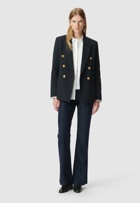 Vrouw met lang blond haar, gekleed in een zwarte dubbelgeknoopte blazer met gouden knopen, wit overhemd, donker uitlopende spijkerbroek en zwarte schoenen tegen een grijze achtergrond.