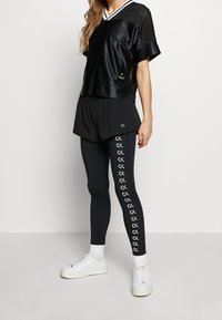 Musta urheilullinen toppi valkoisilla yksityiskohdilla, yhdistetty mustiin shortseihin ja legginsseihin, joissa on logokuvio; valkoiset lenkkarit täydentävät asukokonaisuuden.