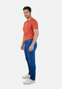 Camiseta roja de manga corta, jeans ajustados azules y zapatillas blancas. La camiseta presenta un pequeño logo en el pecho, proporcionando un aspecto casual.