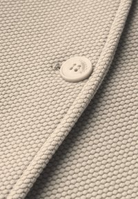 Lichtbeige stof met een gestructureerd breipatroon, voorzien van een ronde, witte knop met vier gaten en subtiele branding op het oppervlak.