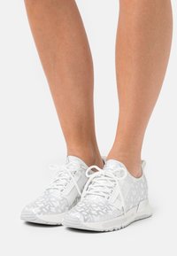 Person in weißen und grauen gemusterten Sneakers mit weißen Schnürsenkeln, die vor einem schlichten weißen Hintergrund stehen.