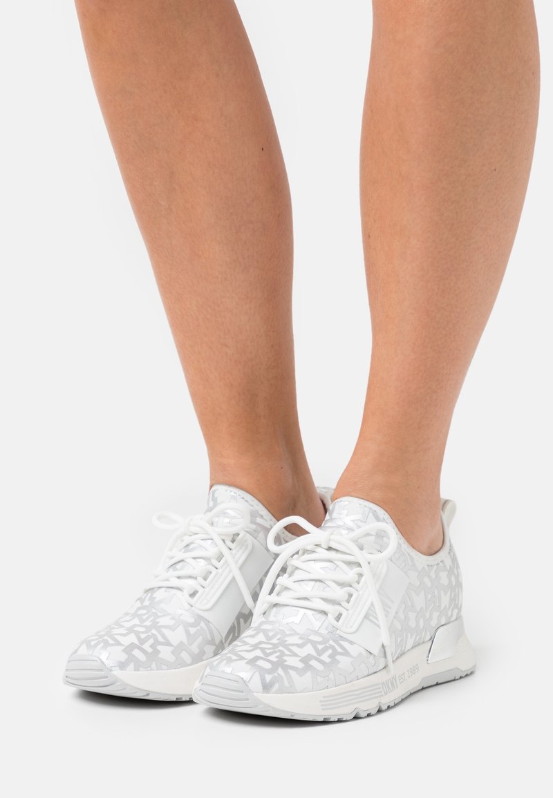Person in weißen und grauen gemusterten Sneakers mit weißen Schnürsenkeln, die vor einem schlichten weißen Hintergrund stehen.
