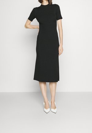Robe midi noire à côtes avec des manches courtes, un col haut et une coupe droite. Associée à des escarpins blancs à bout pointu. Texture lisse sur l'ensemble.