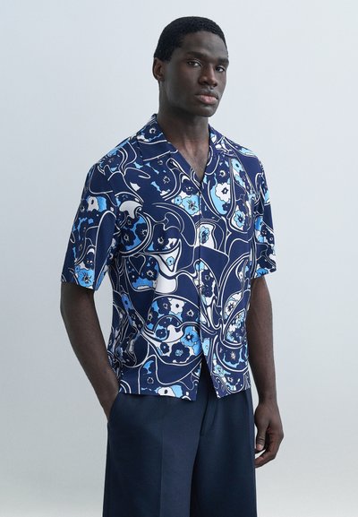 Jeune homme debout, les mains dans les poches, portant une chemise à manches courtes bleue à motifs floraux et un pantalon sombre, sur un fond uni.