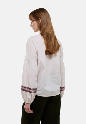 Blouse blanche avec des manches bouffantes et des accents brodés rouges aux poignets, présentant un col rond simple et une texture de tissu lisse.