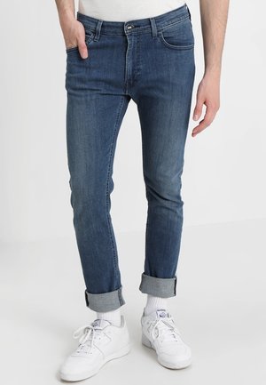 Man draagt opgerolde blauwe slim-fit jeans, witte sokken en witte Reebok-sneakers, staand met linkerhand in de zak tegen een witte achtergrond.