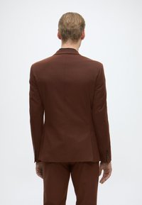 Bruin maatpak met een getailleerd achterontwerp. Glad stof, enkele split en zwarte knoopaccenten op de mouwen. Eenvoudige en moderne esthetiek.