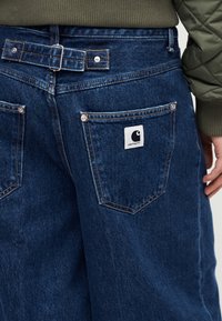 Άτομο που φοράει μπλε τζιν παντελόνι με ρυθμιζόμενο πλευρικό πόρπη και λογότυπο Carhartt στην πίσω τσέπη, συνδυασμένο με χακί καπιτονέ μπουφάν.