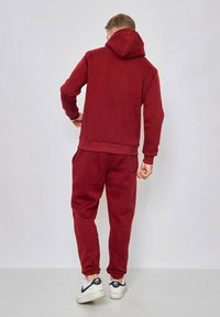 Haut à capuche rouge et pantalons de survêtement assortis en tissu doux. Présente une capuche avec cordon de serrage, des poignets côtelés et une taille élastiquée. Des baskets blanches complètent l'ensemble.