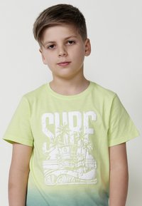 Světle zelené bavlněné tričko s gradientním designem, se bílou grafikou "SURF" a ilustrací palmy. Kulatý výstřih a krátké rukávy.