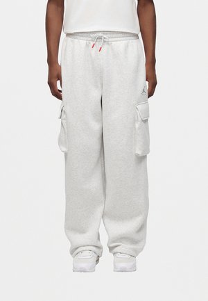 Persona che indossa pantaloni cargo da ginnastica grigio chiaro con vita regolabile e sneakers bianche, in piedi davanti a uno sfondo semplice.