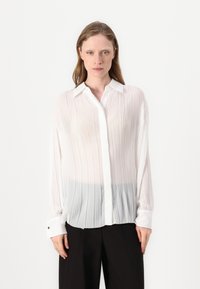 Blusa bianca plissettata con texture trasparente integrata, colletto a camicia e pannello inferiore grigio a contrasto. Maniche lunghe con dettagli ai polsini.