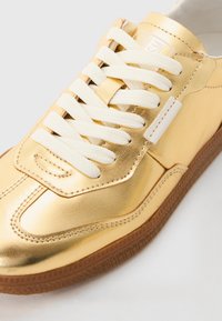 Arany fémes sneaker fehér fűzőkkel és gumitalppal. Simább textúrával, varrott részletekkel és finom logóval a nyelvén.