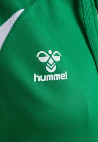 Tissu de sport vert avec logo blanc Hummel, présentant un graphisme stylisé d'abeille et le nom de la marque en dessous, à côté d'un panneau en mesh blanc.