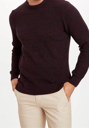Homme portant un pull texturé bordeaux foncé et un pantalon beige clair, les mains dans les poches, sur un fond uni.