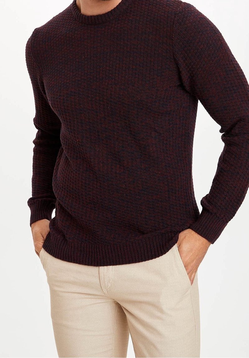 Homme portant un pull texturé bordeaux foncé et un pantalon beige clair, les mains dans les poches, sur un fond uni.