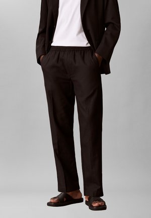 Mann trägt schwarze, locker sitzende Hose mit elastischem Bund, schwarzen Blazer, weißes T-Shirt und schwarze Slip-On-Sandalen vor grauem Hintergrund.