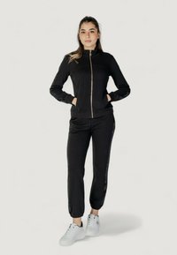 Giacca nera con zip, colletto alto e maniche a polsino, abbinata a pantaloni coordinati con un accento glitterato laterale. Indossata con sneakers bianche.