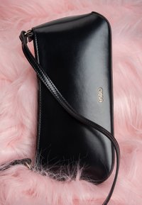 Petit sac à main Estro en cuir noir avec une sangle, posé sur un fond de fausse fourrure rose douce.