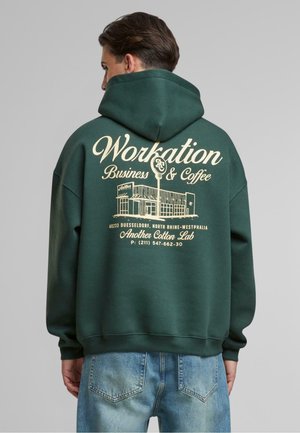 Green hoodie with a large light beige graphic on the back featuring "Workation Business & Coffee," text, and a building illustration.