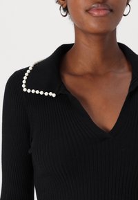 Svart ribbad topp med V-ringning och en krage prydd med vita pärlkantade detaljer längs kanten. Mjuk textur och åtsittande design.