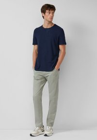 Jonge man staat met zijn handen in de zakken, draagt een marineblauw overhemd met korte mouwen, lichtgrijze broek en witte sneakers, tegen een effen achtergrond.