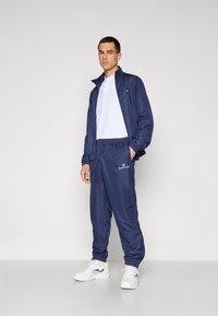 Marineblaues Sport-Trainer-Set mit Reißverschlussjacke und Hose mit elastischem Bund. Weiße T-Shirt darunter, getragen mit weißen Sneakern.
