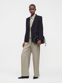 Femme portant un blazer noir croisé, chemise rayée, pantalon beige à jambes larges, chaussures noires pointues, tenant un sac à main en cuir noir.