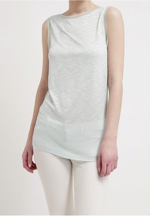 Ärmelloses, hellgraues Strickoberteil kombiniert mit eng anliegenden, cremefarbenen Hosen, getragen von einer stehenden Person vor schlichtem Hintergrund.