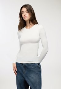 Maglietta bianca a maniche lunghe, aderente, con colletto rotondo, abbinata a jeans in denim larghi. Texture liscia, design minimale e colore neutro.