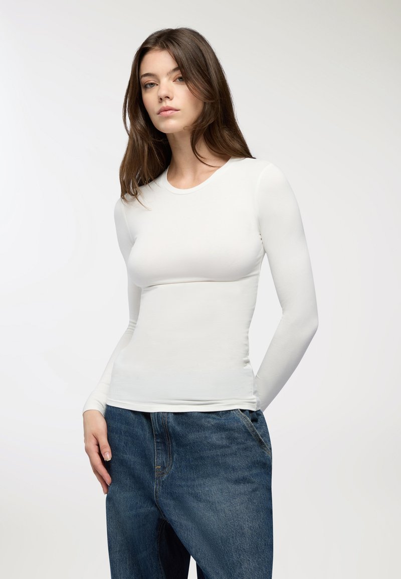 Maglietta bianca a maniche lunghe, aderente, con colletto rotondo, abbinata a jeans in denim larghi. Texture liscia, design minimale e colore neutro.