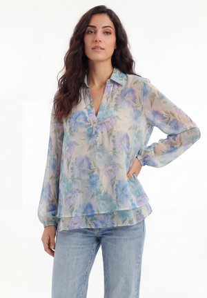 Vrouw met lang donker haar, gekleed in een doorschijnende blouse met bloemenmotief in pastelblauw en paars, gecombineerd met lichtblauwe spijkerbroek, staand met hand op heup.