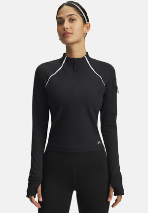 Vrouw in een zwart sportoverhemd met lange mouwen, witte piping, duimopeningen, een ritszak op de mouw en een kwart-rits halslijn.