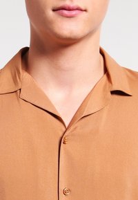 Brun kortärmad skjorta gjord av ett mjukt tyg. Den har en skjortkrage och tre knappar, med en subtil lyster och en skräddarsydd passform.