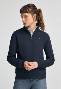 Felpa con zip colore navy realizzata in tessuto morbido, caratterizzata da un collo alto, polsini a costine e un piccolo logo rosso sul petto.