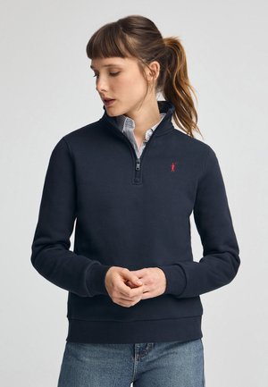 Felpa con zip colore navy realizzata in tessuto morbido, caratterizzata da un collo alto, polsini a costine e un piccolo logo rosso sul petto.