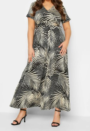 Kobieta ubrana w czarno-beżową sukienkę kopertową z wzorem liści palmy, z krótkimi rękawami i czarnymi szpilkami z paskiem wokół kostki, stojąca na tle białego tła.