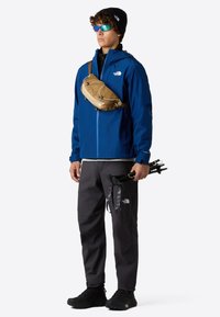 Modrá bunda, černé kalhoty, hnědá kabelka přes rameno, černé boty a sluneční brýle. Ruce držící trekingové hole. Materiál vypadá jako voděodolný.