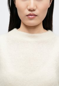 Kreemikvalge kootud kampsun soonikkoelise kaelusega, pehme tekstuuri ja lõdva lõikega, nähtav ülaosa rindkere piirkonnas.