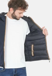 Giubbotto imbottito blu navy con cappuccio, dotato di una tasca interna con zip e un accento a righe. Tessuto liscio, trapuntato e materiale leggero.