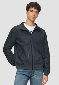 Marine blauw windjack met een ritssluiting aan de voorkant, een staande kraag en knoopzakken; gladde textuur en elastische manchetten. Gecombineerd met lichte jeans.