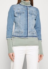 Blekt blå denimjacka med fransad nederkant, med två bröstfickor och dragkedjestängning, bära över en ljusgrön långärmad topp.