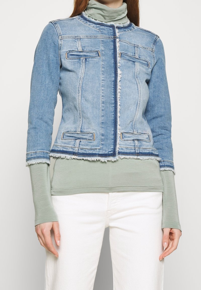 Blekt blå denimjacka med fransad nederkant, med två bröstfickor och dragkedjestängning, bära över en ljusgrön långärmad topp.